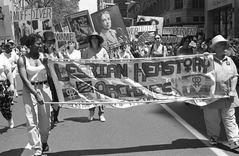 Two lesbians carry the Lesbian Herstory Archives banner in a march, with a crowd of lesbians behind them carrying signs like "Muff-Diver," "Invert," "Mannish," and "Dyke."