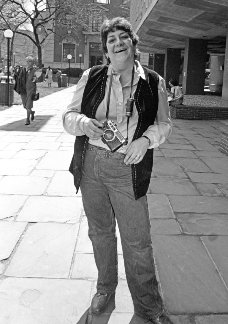 Black and white photo of Madeline Davis wearing a long-sleeved shirt, a dark vest, and pants standing on the sidewalk on a sunny day smiling into the camera.