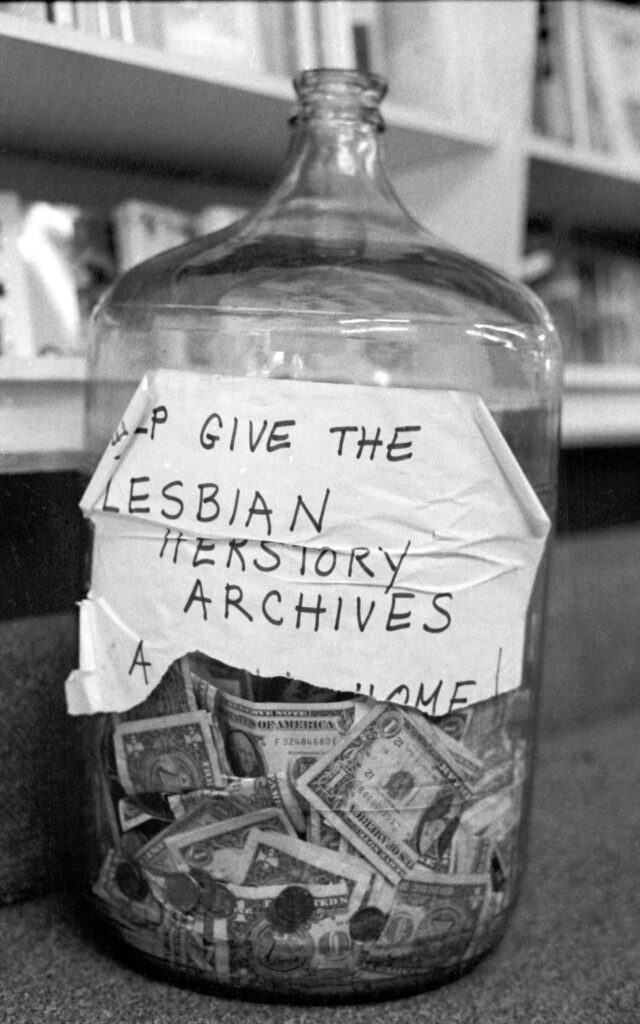A large, clear glass bottle with many dollar bills and coins visible at the bottom and a torn sign taped to it that reads, Help Give the Lesbian Herstory Archives A New Home.
