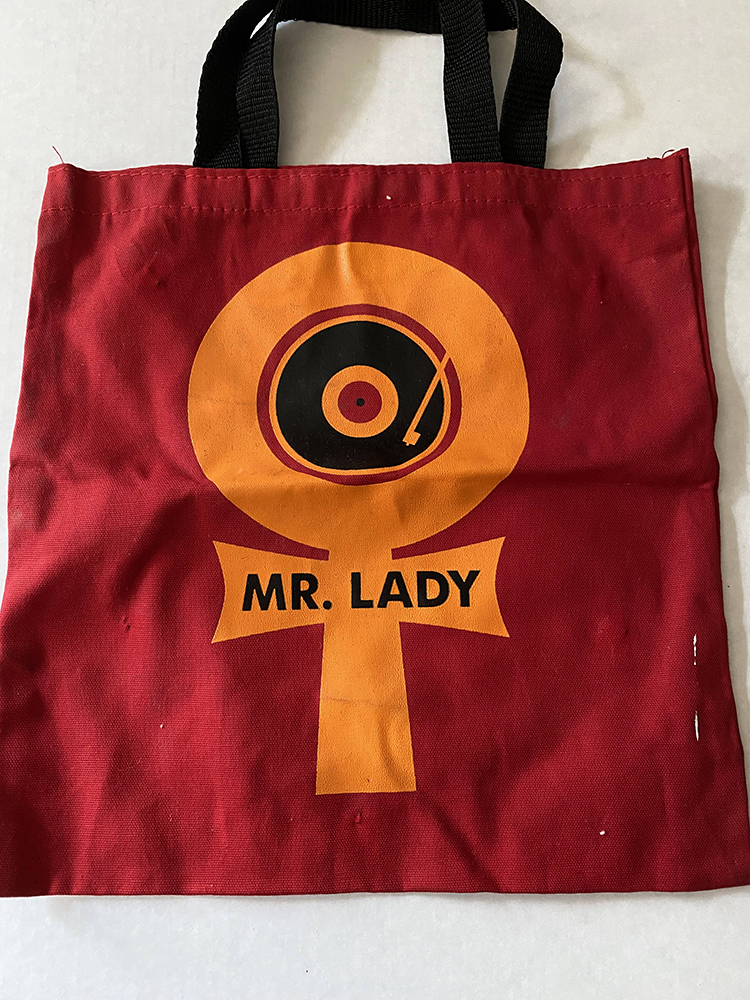 A dark red, rectangular canvas tote bag with black nylon handles sites on a white table. On the tote back is a large orange and black logo, made up of a female symbol with a "MR. LADY" printed over the cross bar at the bottom and a graphic representation of a record player at the center of the circular portion at the top.